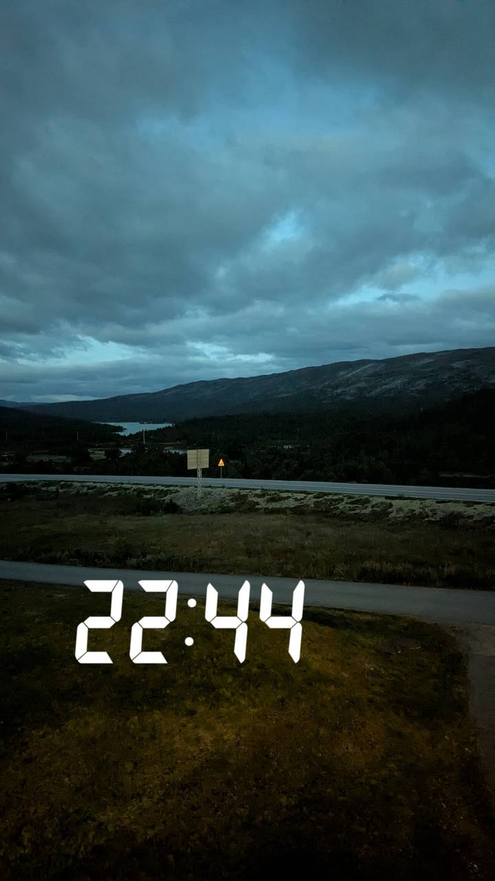 Dim pre-dawn view of a highway and distant lake against dark rolling mountains under a cloudy sky.