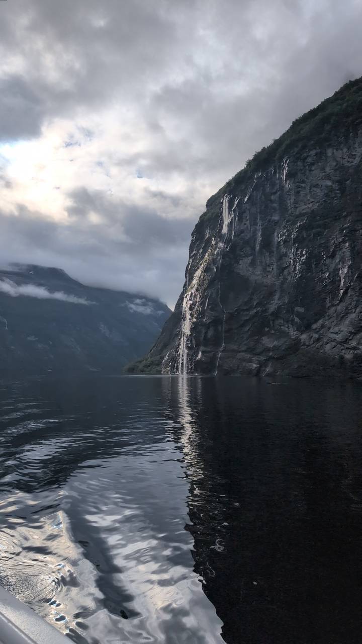 Dramatic waterfall plunges down a sheer cliff into a calm fjord framed by misty mountains.