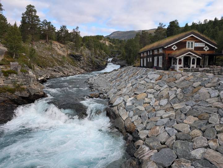 Rushing turquoise river flows past a stone-lined bank and a turf-roof cabin in a mountain valley.