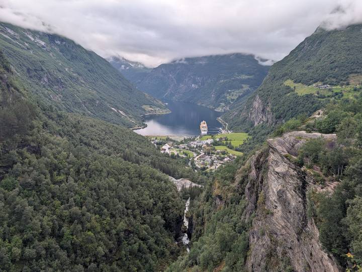 Panoramic view over Geirangerfjord with cruise ship, village, and lush green cliffs under low clouds.