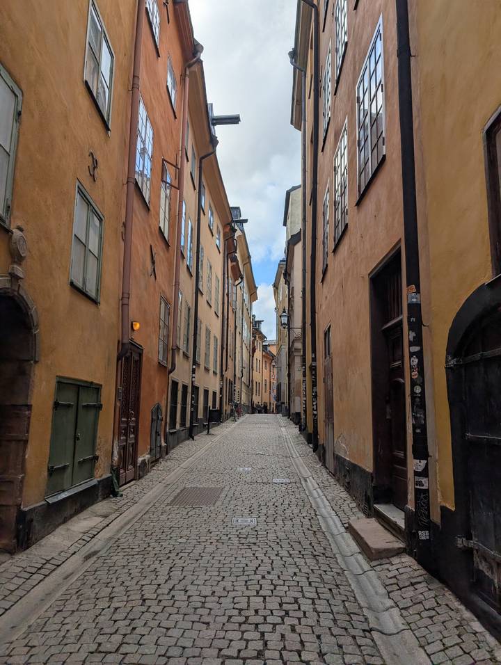 Cobbled narrow lane lined with warm-hued historic buildings in Stockholm’s old town under patchy clouds.