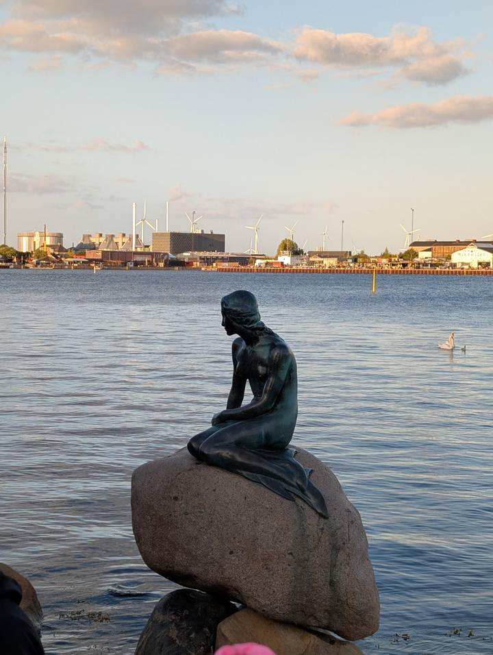 Iconic Little Mermaid statue gazes over the harbor with industrial skyline and wind turbines behind.