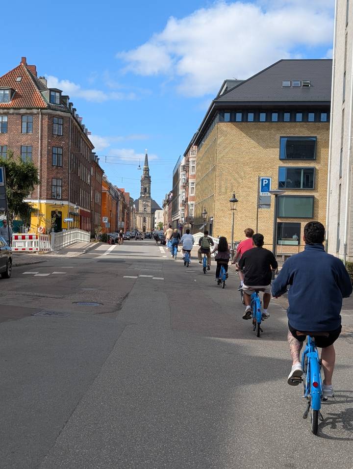 Line of cyclists rides down a Copenhagen street toward a historic tower on a cool, bright day.