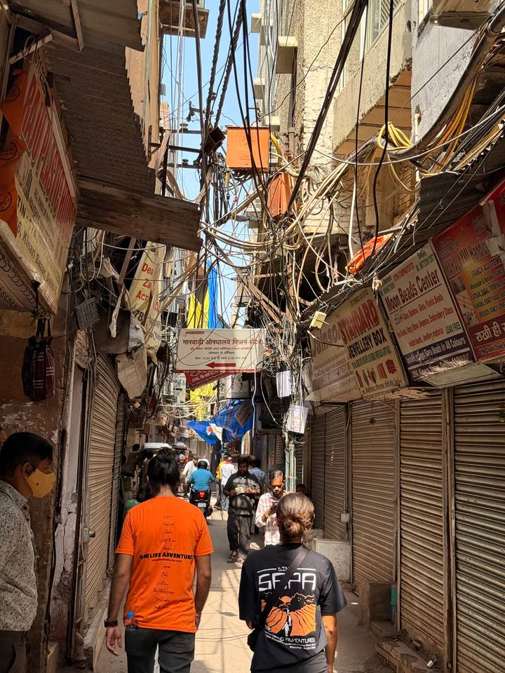 Tangled electrical wires hang above a bustling narrow lane filled with signage and shoppers.