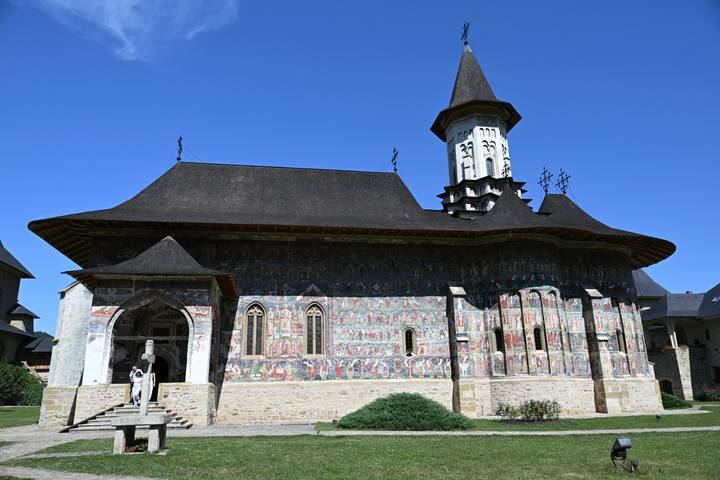 Voroneț Monastery with dark shingled roof and colourful exterior fresco panels under a blue sky.