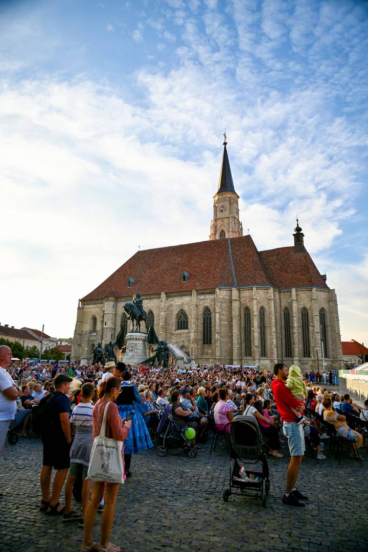 Large Gothic church with a mounted statue in front, surrounded by a dense crowd in a historic square under a blue sky