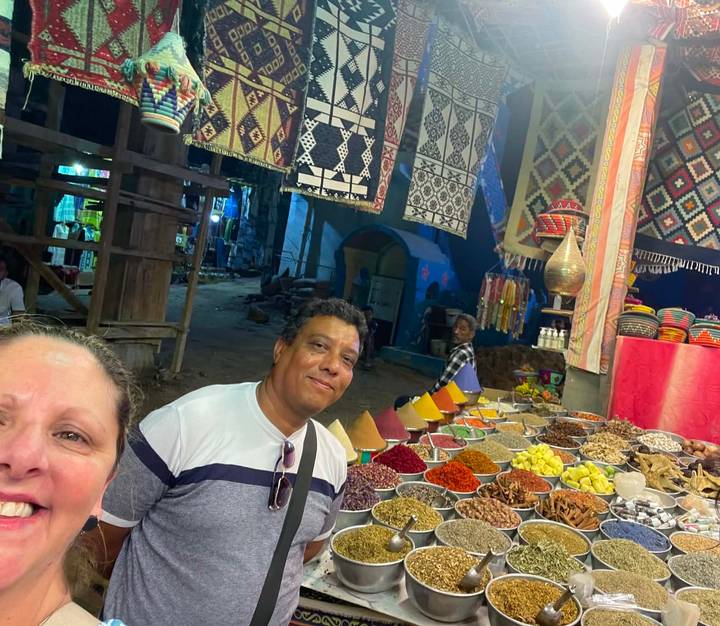 Tourists smile in front of a colorful spice and herb stall at a bustling market.