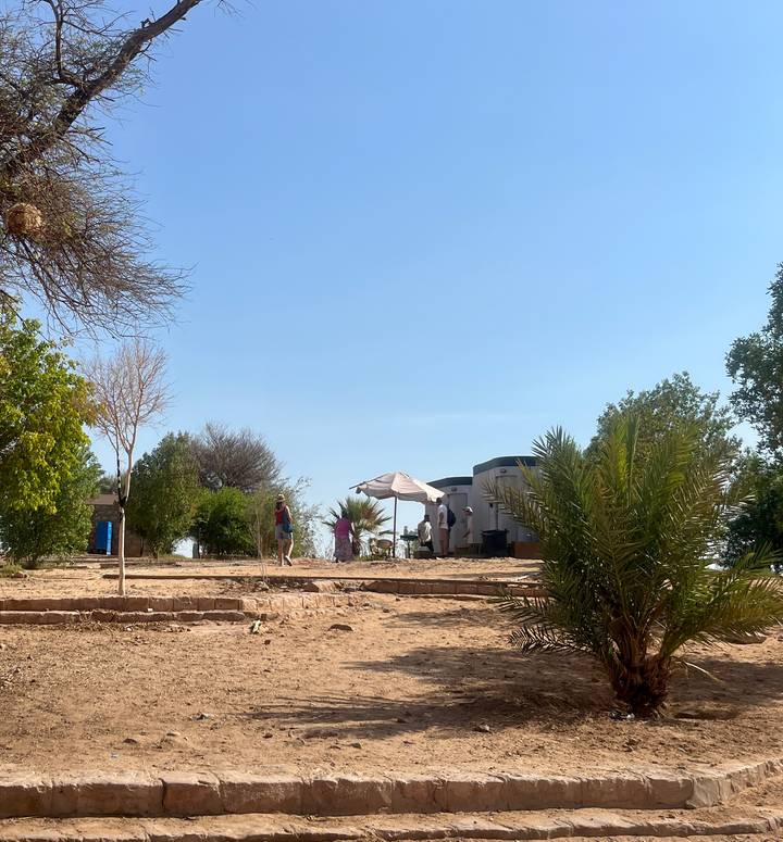 Small group relaxes near simple huts and palm trees on a sandy riverside terrace.