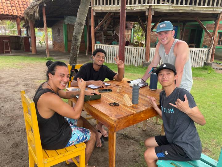 Group of surfers relax at an outdoor wooden table at a beach lodge surrounded by palm trees.