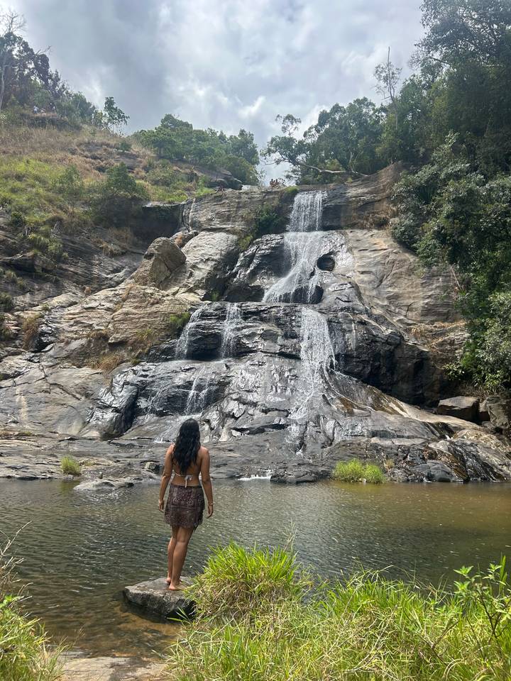A woman stands before a cascading jungle waterfall tumbling over dark rock.
