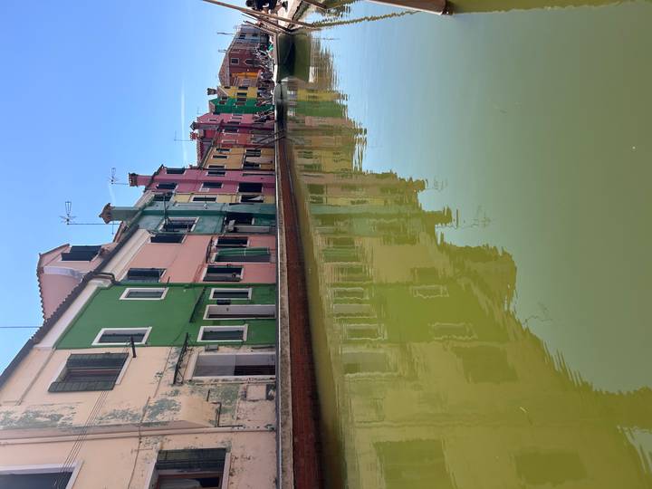 Colorful pastel houses of Burano reflected in a quiet canal under clear blue sky.