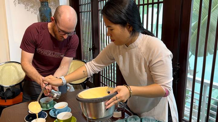 A barista teaches a visitor how to pour Vietnamese egg coffee in a cozy café setting.