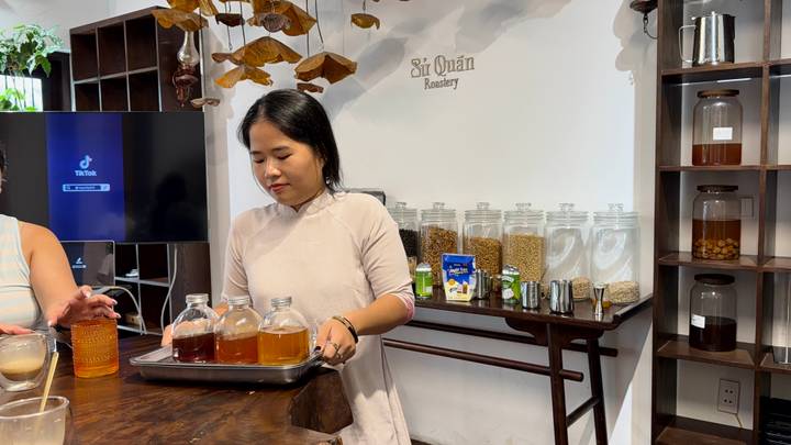 A host carries a tray of tea and coffee samples across a tasting room lined with glass jars of beans.