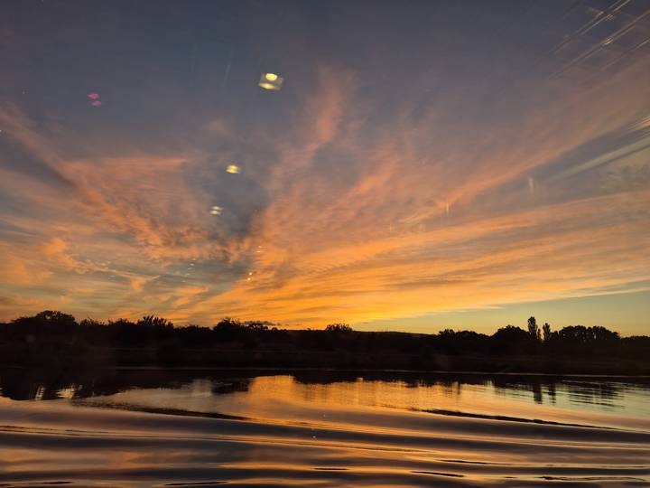 Orange and pink clouds are reflected on a calm river at dusk, viewed through a window.