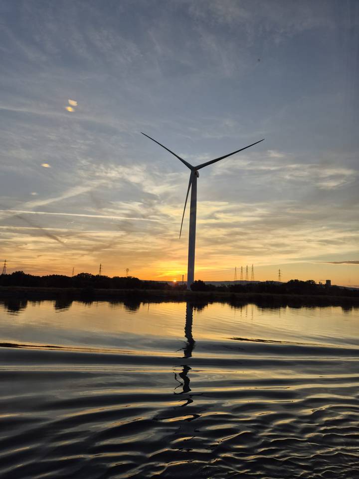 A tall wind turbine stands beside a reflective river during a golden sunset.