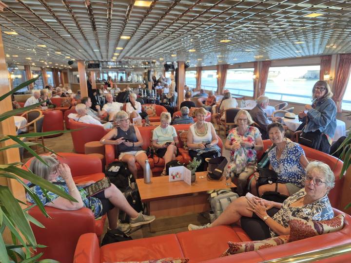 Dozens of cruise passengers relax in a spacious lounge filled with seating and large windows.