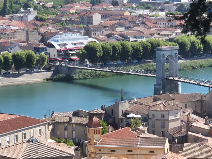 A historic suspension bridge spans a turquoise river flanked by tree-lined streets and tiled rooftops.