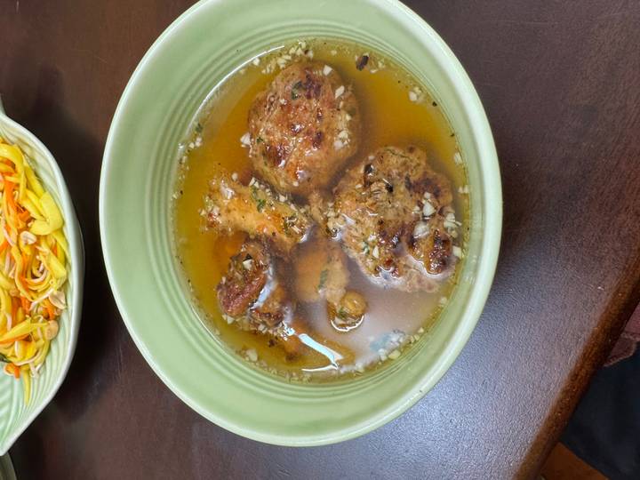 Bowl of Vietnamese broth with grilled meat patties and herbs on wooden table