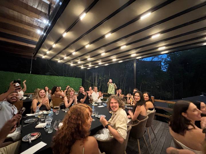 Large group of diners clap and celebrate around a long outdoor table at night.