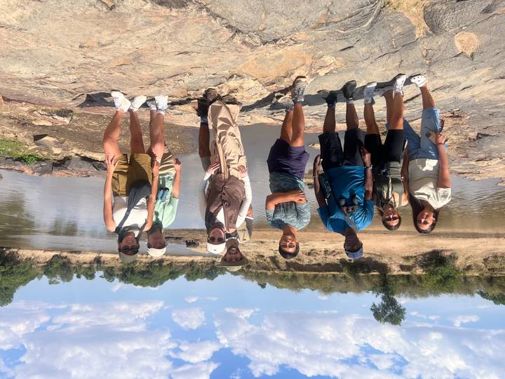 Tourists pose on rocky ground beside a wide river under a blue sky in the savanna.