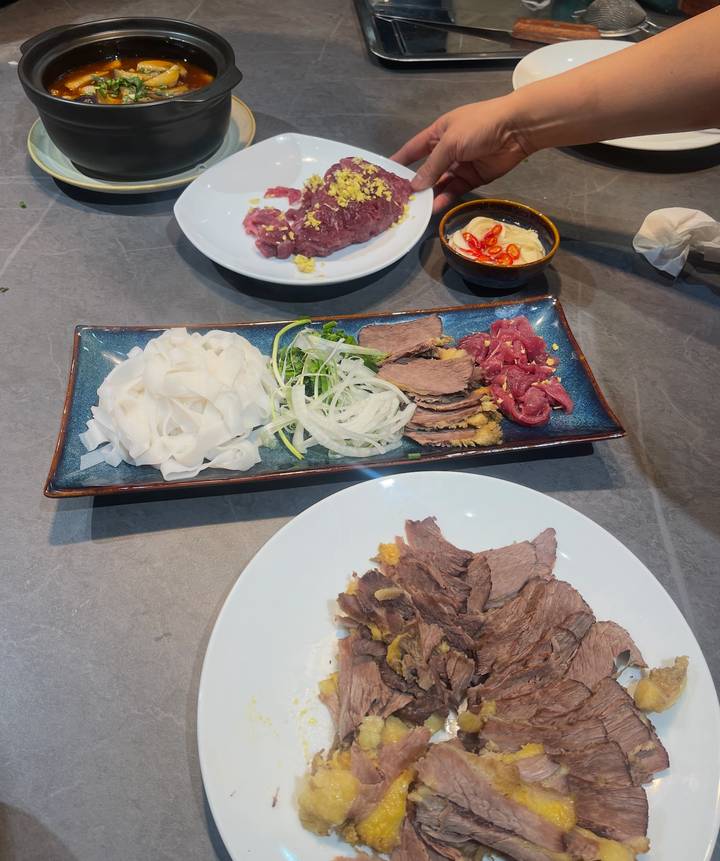 Rectangular platter with pho ingredients: rice noodles, sliced onion, cooked beef slices and raw beef on a grey tabletop.
