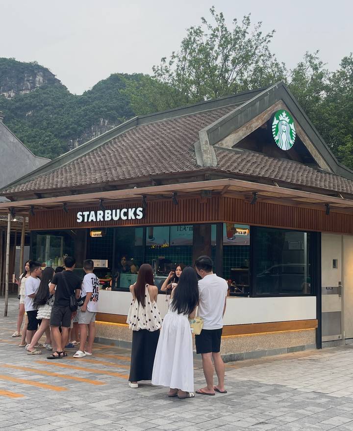 Customers queue outside a modern wooden Starbucks kiosk set against green hills.