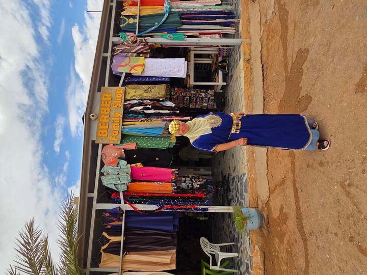 Visitor in traditional blue Berber dress stands outside a colorful family shop.