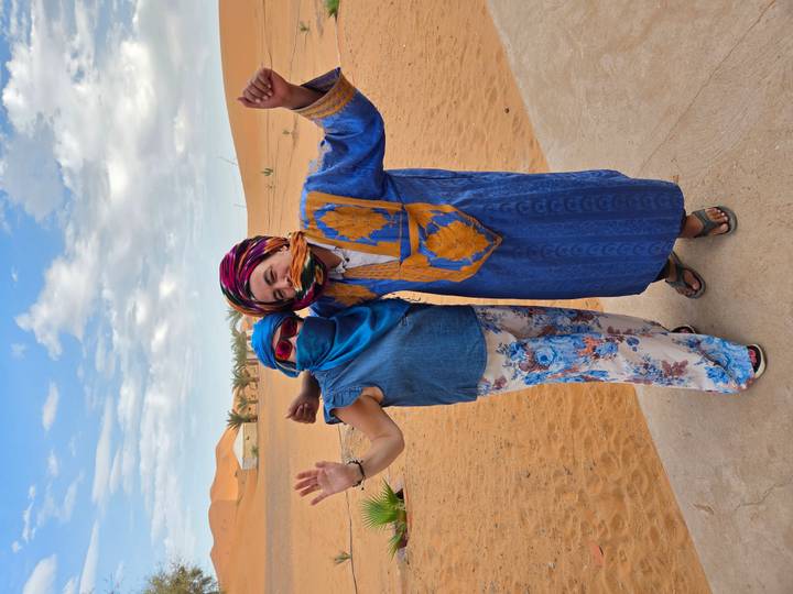 Two travelers in colorful scarves pose playfully on the edge of desert dunes.