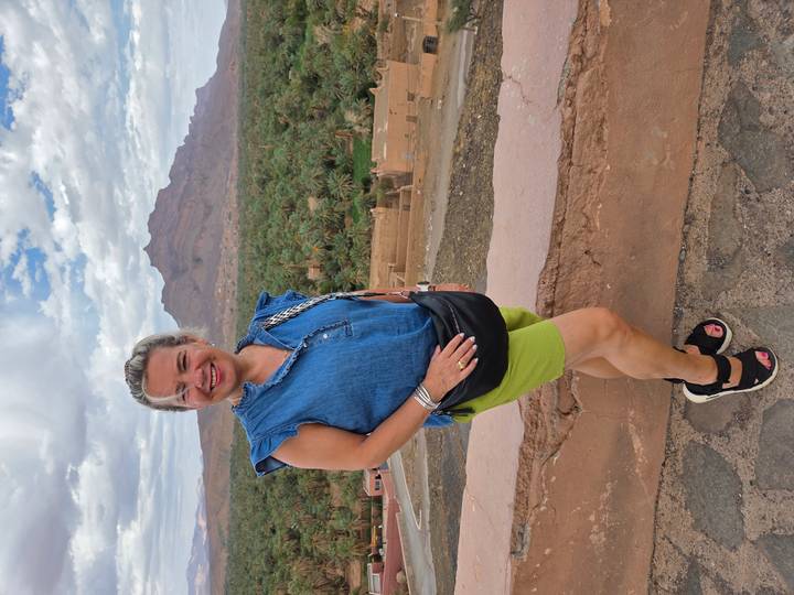Smiling traveler stands before lush oasis of palm trees and a distant rocky peak.