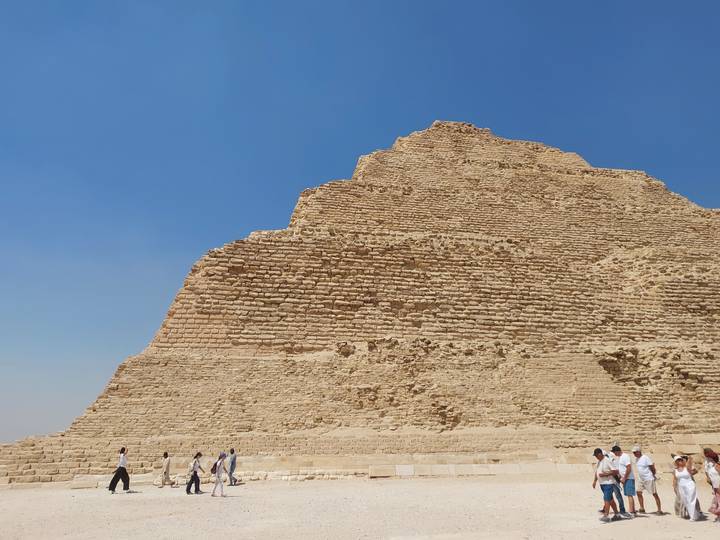 Stepped Pyramid of Djoser in Saqqara with tourists dwarfed at its base under a blue sky.