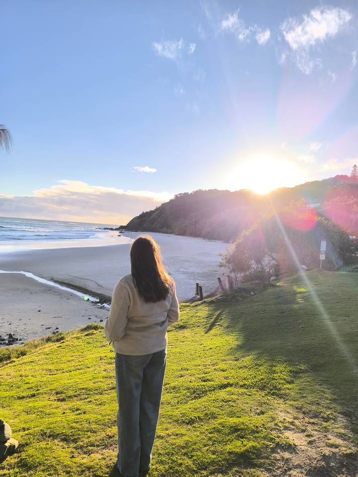 Person gazes at a quiet beach and ocean as the warm golden sun sets behind a tree-covered hill.