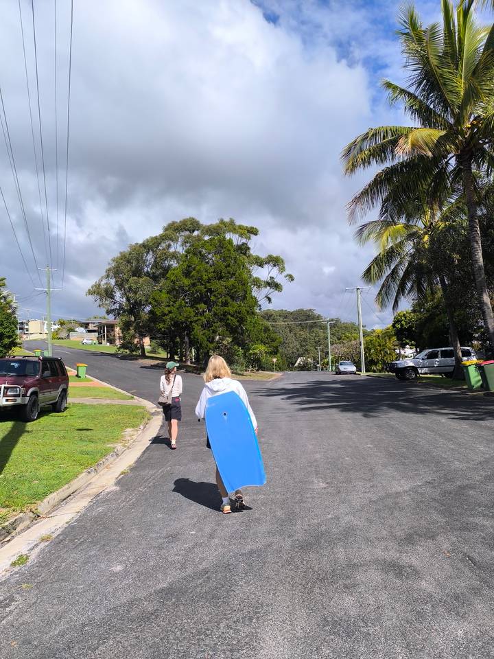 Two beachgoers carry a bodyboard up a quiet residential street beneath gray clouds.