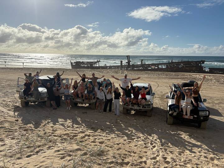 Energetic tour group poses atop three 4WD vehicles on a sandy beach with a famous shipwreck behind.