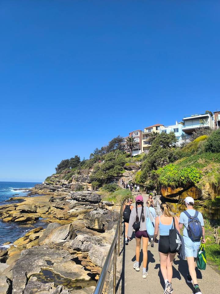 Coastal cliff pathway with walkers and colorful hillside homes overlooking the bright blue sea.