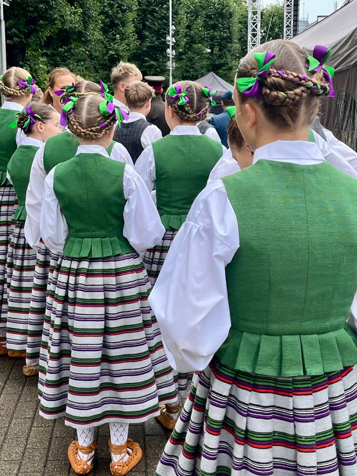 Back view of participants wearing bright green traditional folk vests over striped skirts and white shirts.