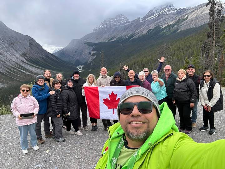 Large tour group poses with Canadian flag for a selfie in front of dramatic Rocky Mountain valley.