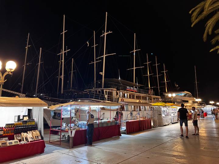 Nighttime market stalls line a marina filled with tall-masted sailing yachts under lamplight.