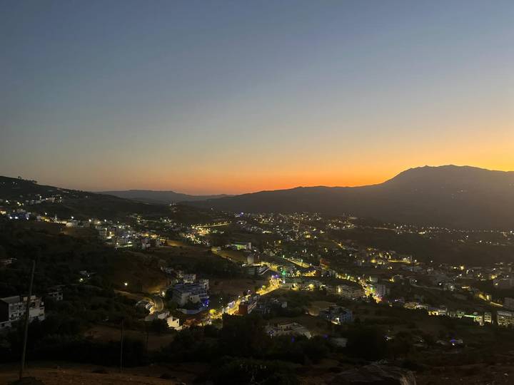 Twilight panorama of a hilly Moroccan town with lights twinkling beneath an orange sky.