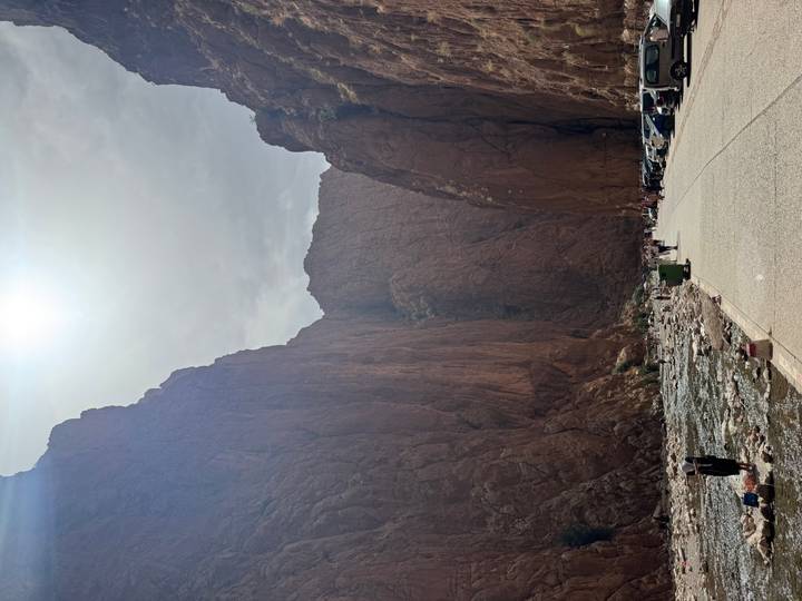 Visitors walk along the narrow floor of Todra Gorge beside a shallow river under towering cliffs.