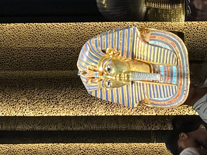 Golden funerary mask of Tutankhamun displayed in a museum case with patterned backdrop.