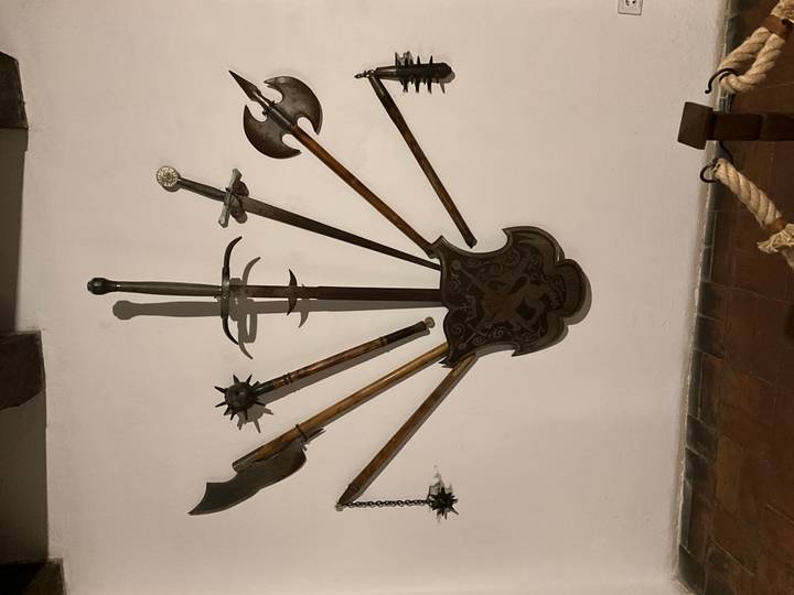 Indoor museum display of medieval weapons arranged around a decorative shield on a white plaster wall.