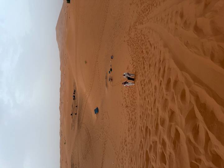 Two travelers climbing up steep sand dunes with scattered desert camp tents visible in the distance.