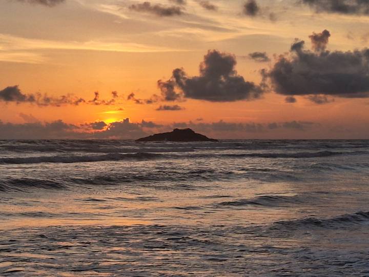 Vibrant orange sunset over the ocean with small rocky islet on the horizon and rippling waves in the foreground.