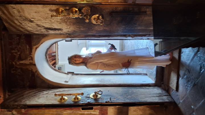 Woman in flowing white outfit stands inside an antique arched doorway of a dimly lit historic building.