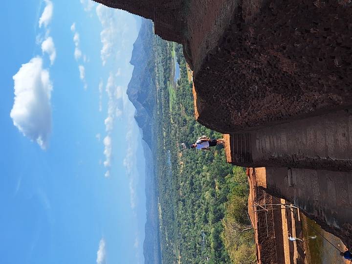 Hiker stands on ancient stone steps overlooking vast green plains and distant mountains from a high rock fortress.