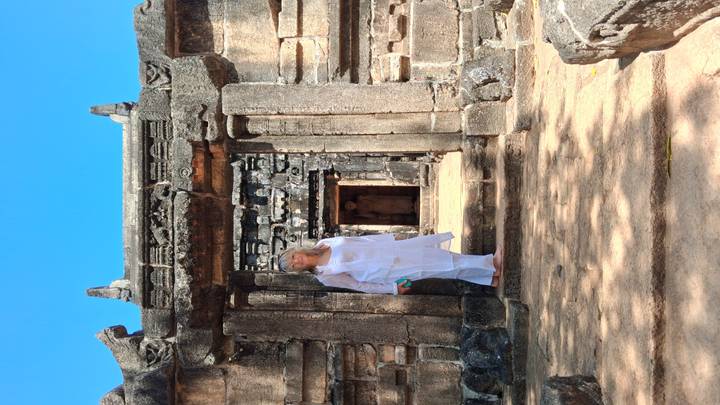 Woman in white attire stands at the stone entrance of an ancient ruin complex under bright blue sky.