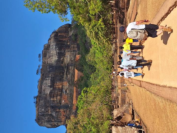 Line of tourists walking along a sandy path toward the massive vertical face of Sigiriya Rock.