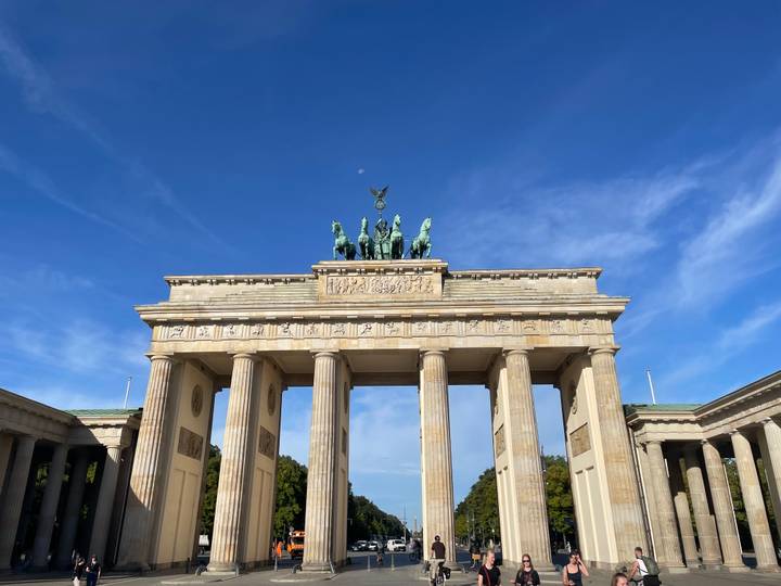 Daytime view of Brandenburg Gate under a clear blue sky, small moon visible above.