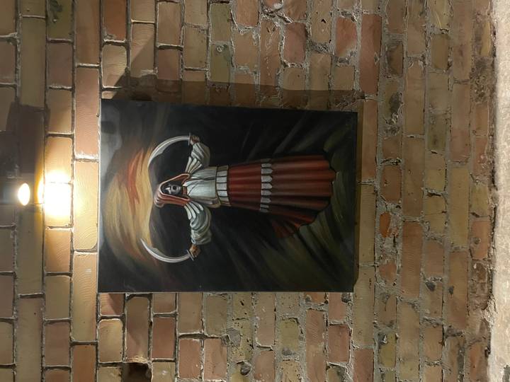 Painting of a hooded figure with scythes displayed on an exposed brick wall under a spotlight.