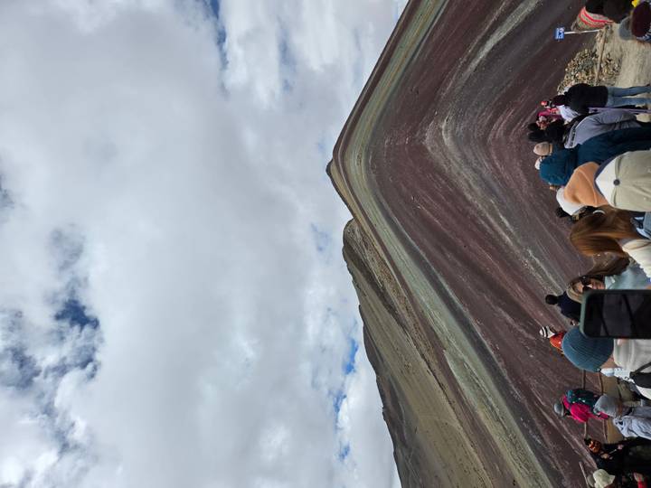 Visitors bundled in jackets hiking along the colorful striations of Rainbow Mountain under a cloudy sky